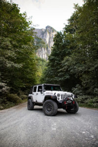 A Jeep Wrangler is parked on road between tall trees, Yosemite's El Capitan wall in California is behind them
